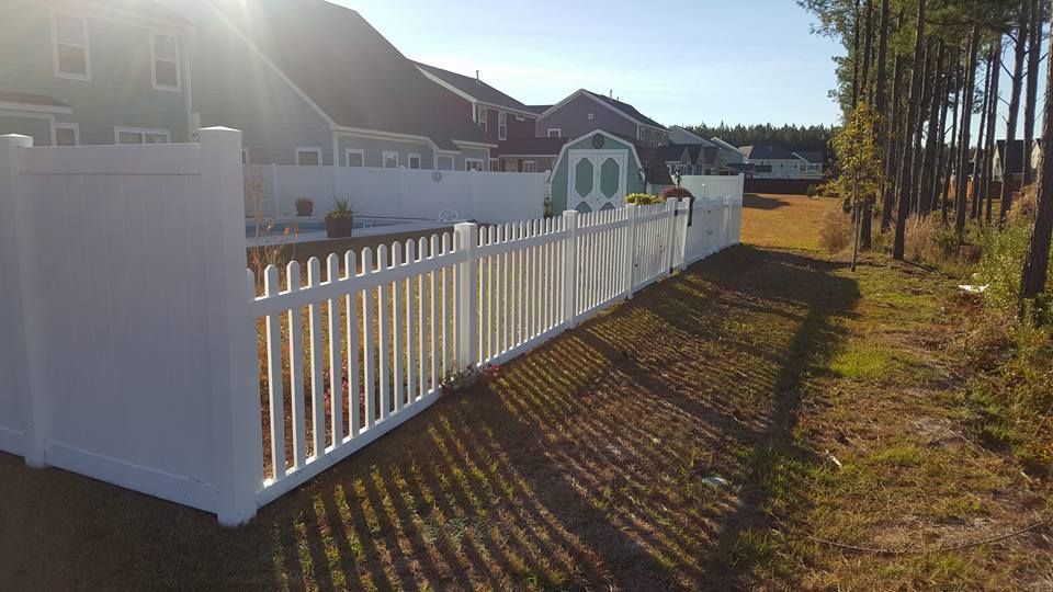 A white picket fence is surrounded by grass and trees in front of a house.