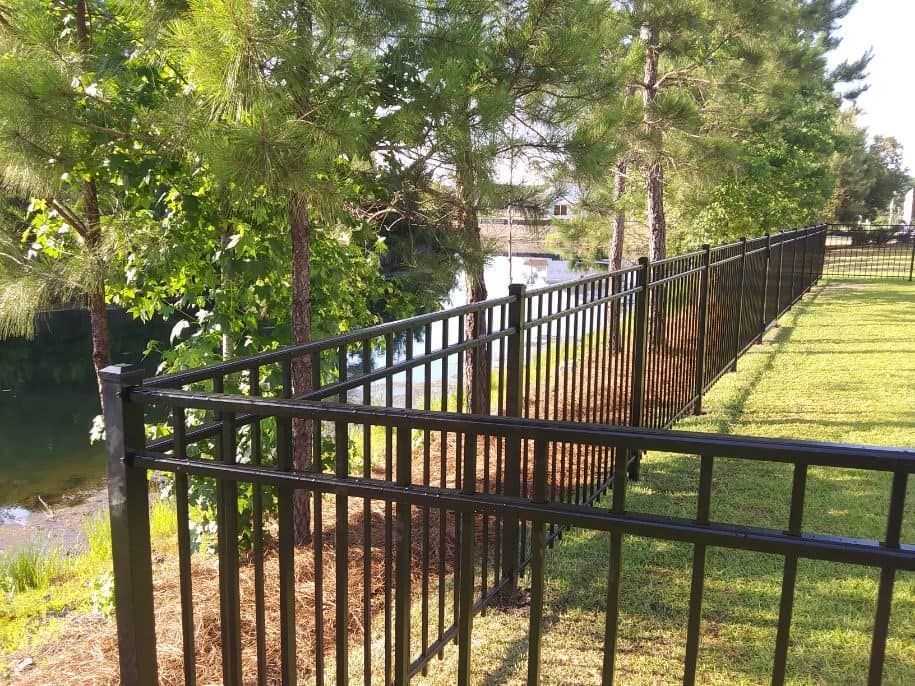 A black fence surrounds a lush green field next to a lake.
