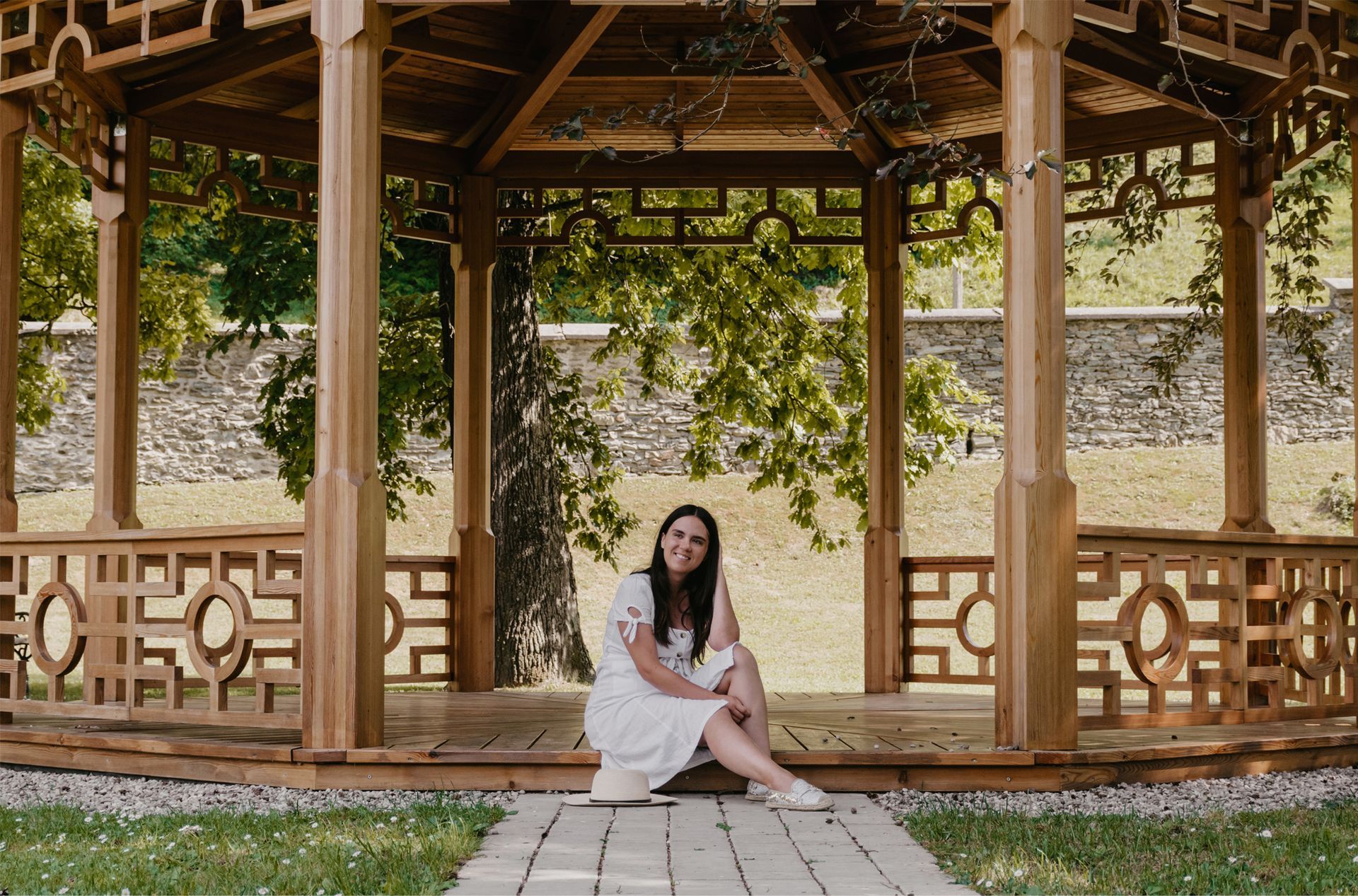 A woman in a white dress is sitting under a wooden gazebo.