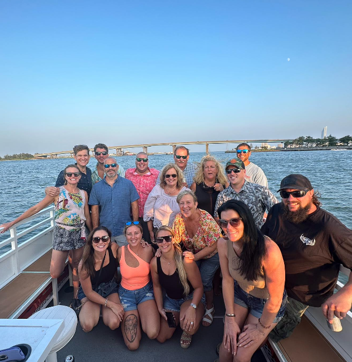 Group of people on a boat with bridge in the background. Smiling faces, sunny day, clear blue sky.