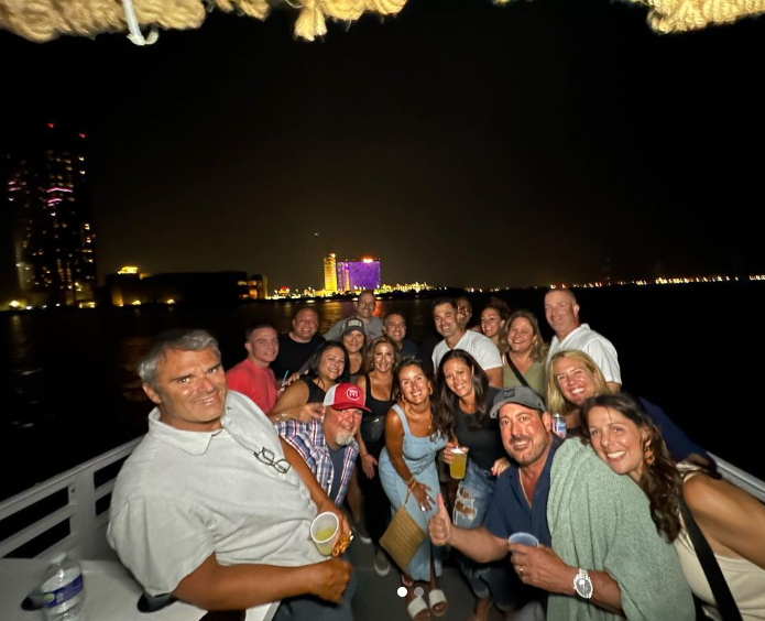 Group of people on a boat at night, city lights in the background. They are smiling, holding drinks.