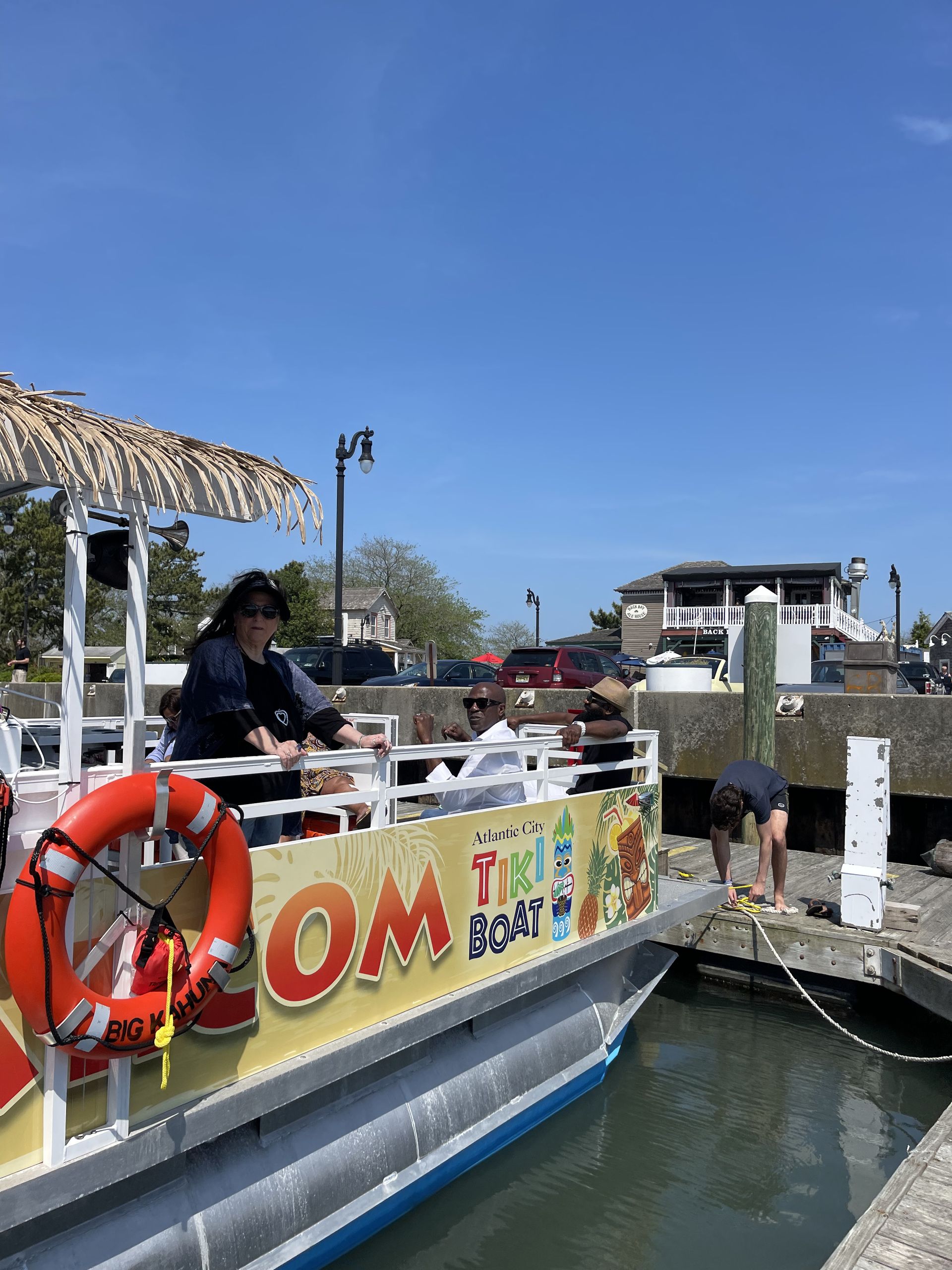 Tiki boat on water, people aboard. A man near dock, blue sky.