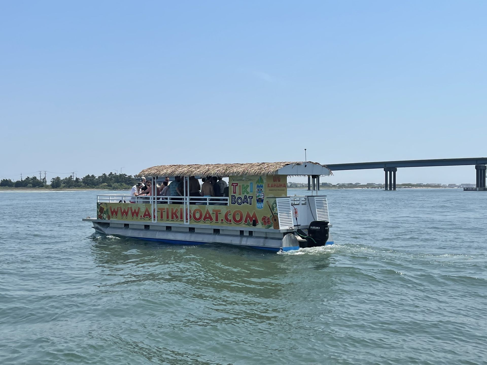 Tiki boat on water with passengers; a bridge and shoreline in the background on a sunny day.