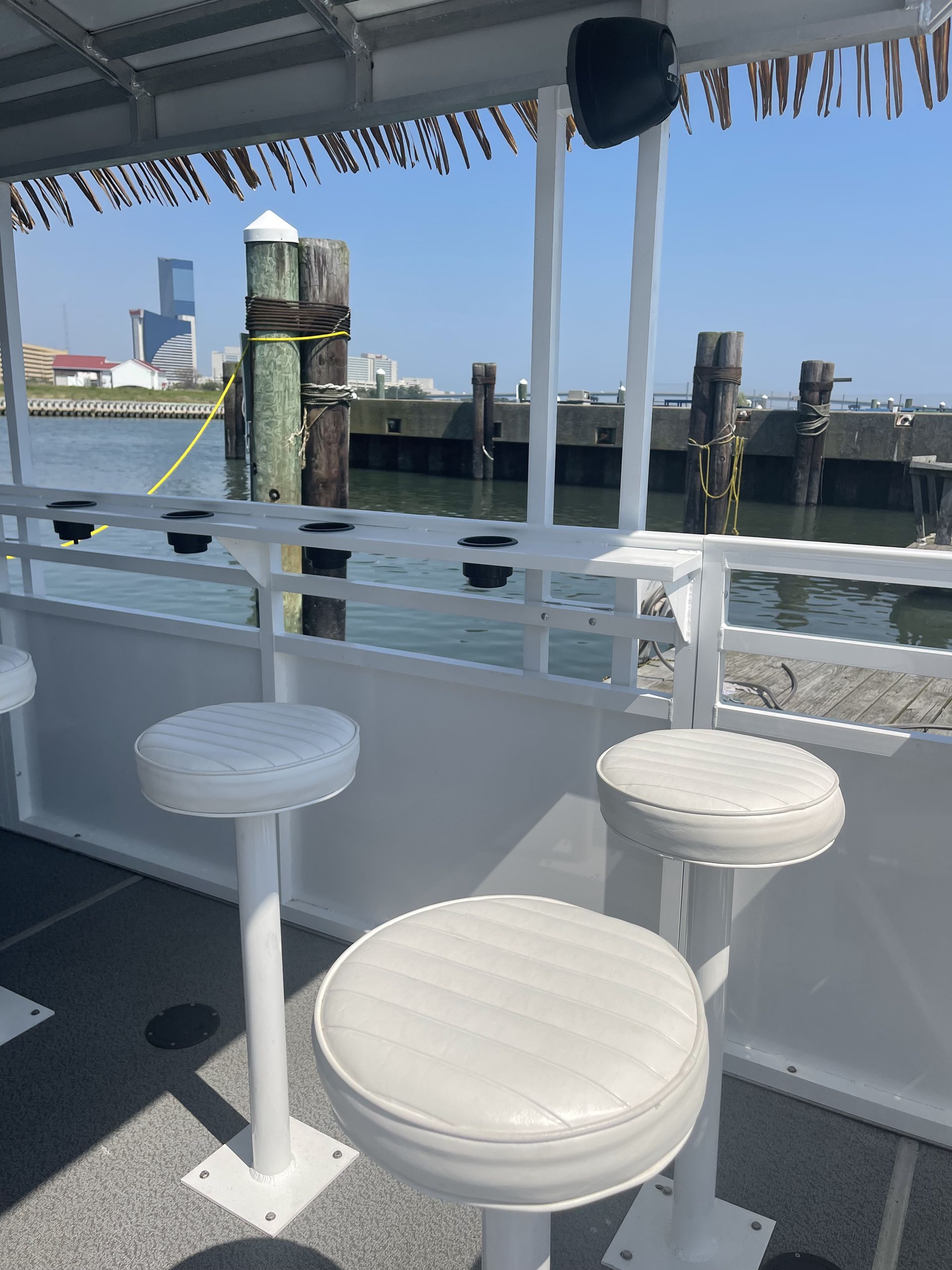 White stools on a boat deck overlooking a pier and water, with a city skyline in the background.