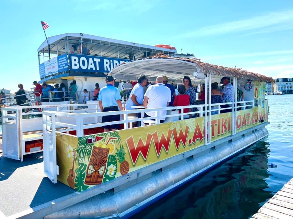Tiki boat with people on the water. Brightly decorated with a thatched roof, in front of a larger boat. Sunny day.