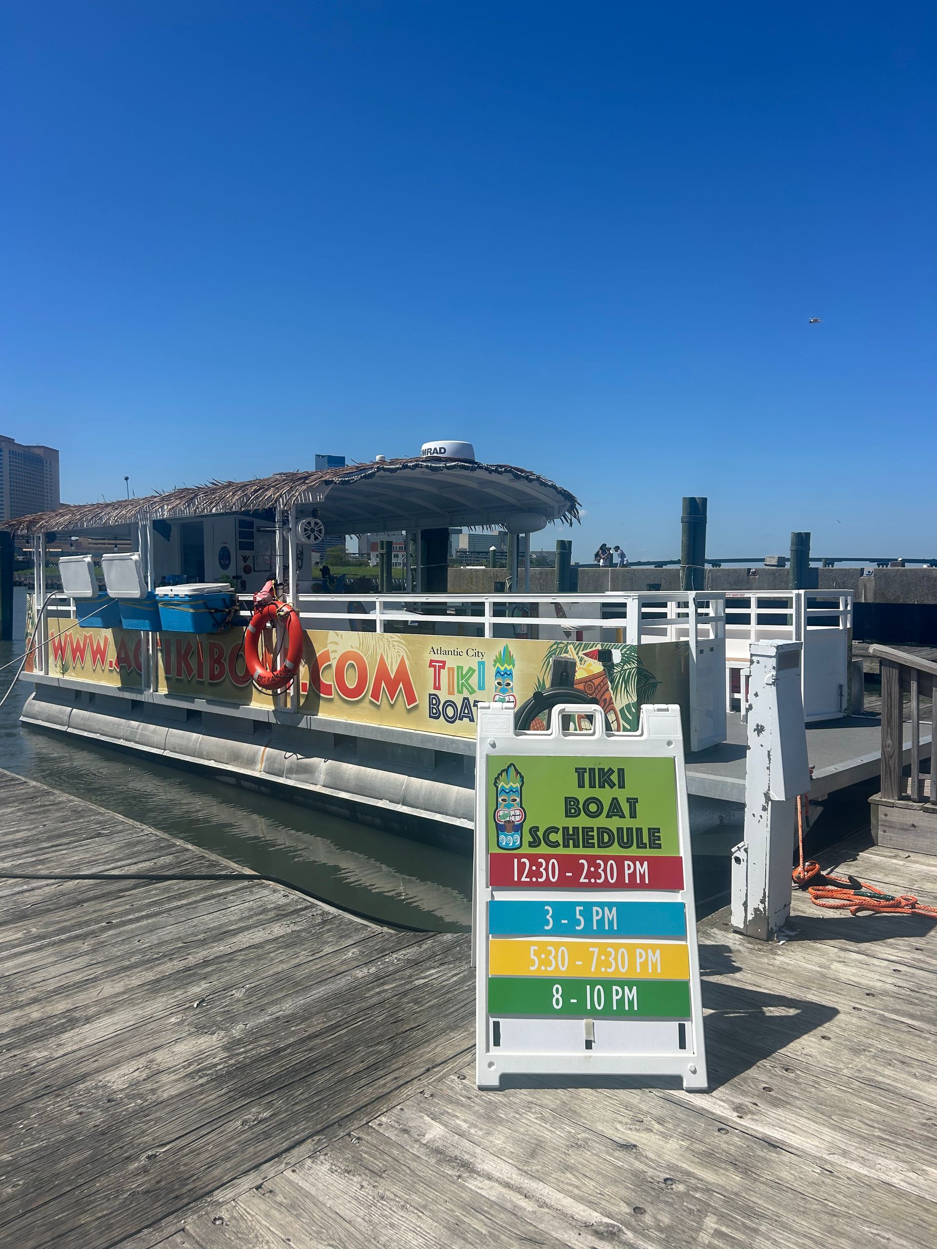 Boat docked on a pier with a sign listing tour times: 1:30 PM, 2:30 PM, 3:30 PM, 4:30 PM. Blue sky.
