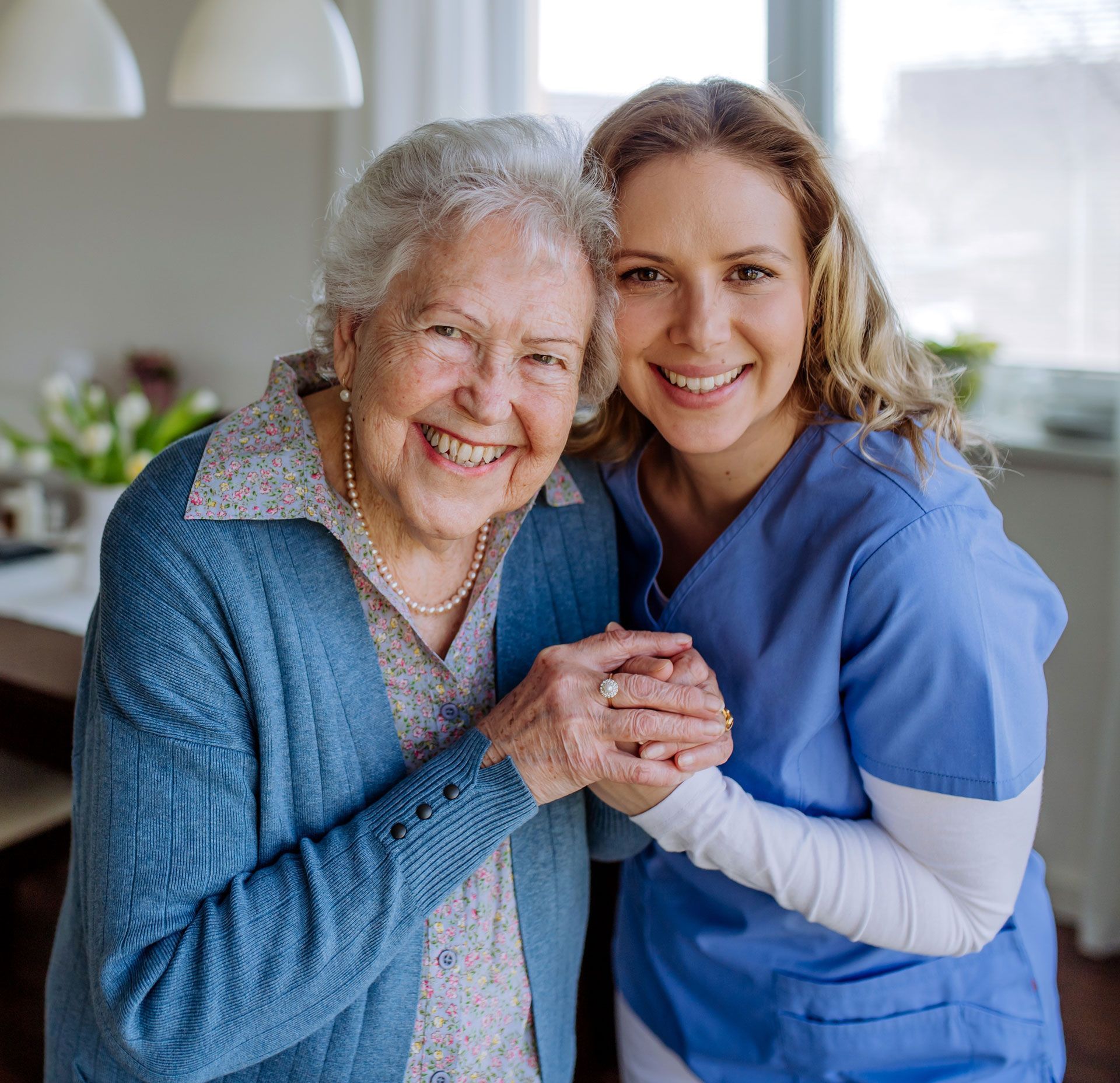 A person in blue medical scrubs holding hands with an older person, both smiling in a bright, indoor setting.