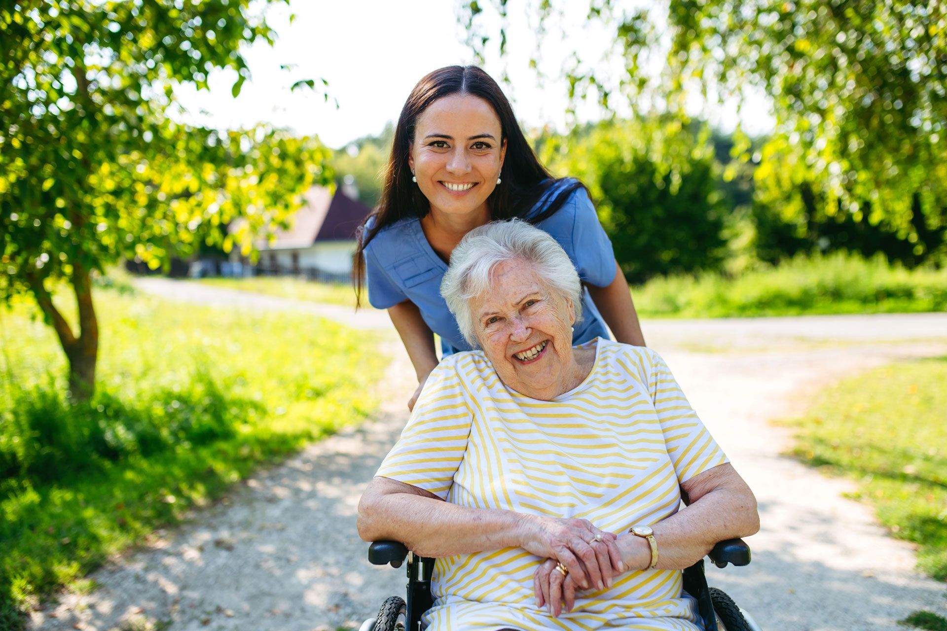 A caregiver in blue scrubs stands behind a smiling person in a wheelchair on a sunny, tree-lined gravel path.