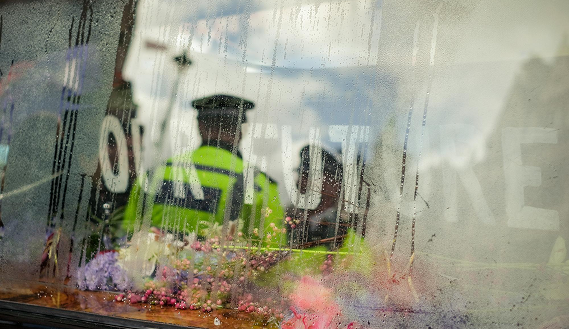 A police officer in a high-visibility yellow vest stands behind a glass window decorated with flowers and the word