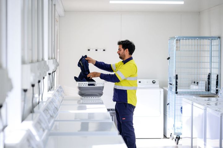 Man in work clothes loading clothes into a washing machine in a well-lit laundry room.
