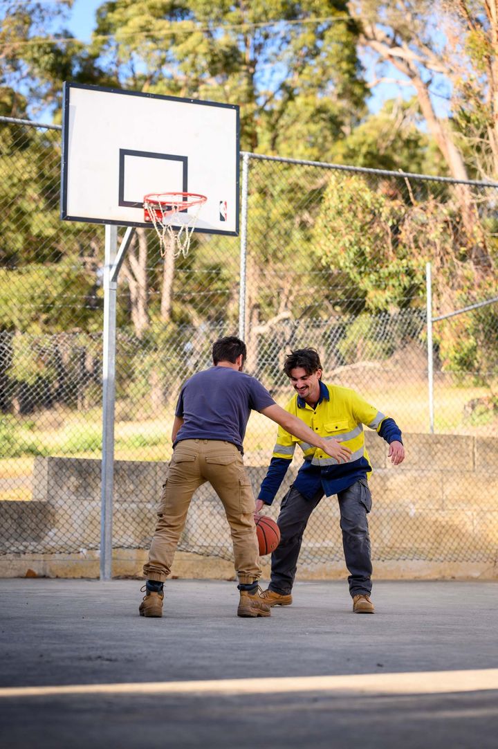 Two men playing basketball on a court. One in work clothes dribbles, the other defends. Hoop and fence in background.