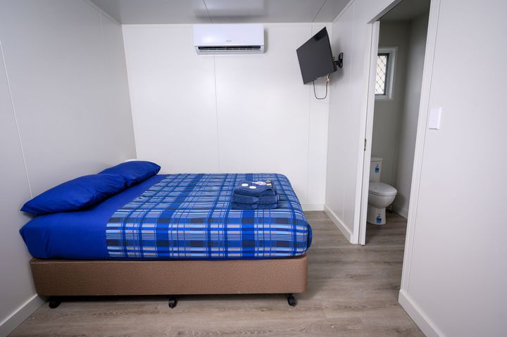 Small, simple worker accommodation bedroom with a bed, air conditioner, TV, and open doorway to a bathroom.