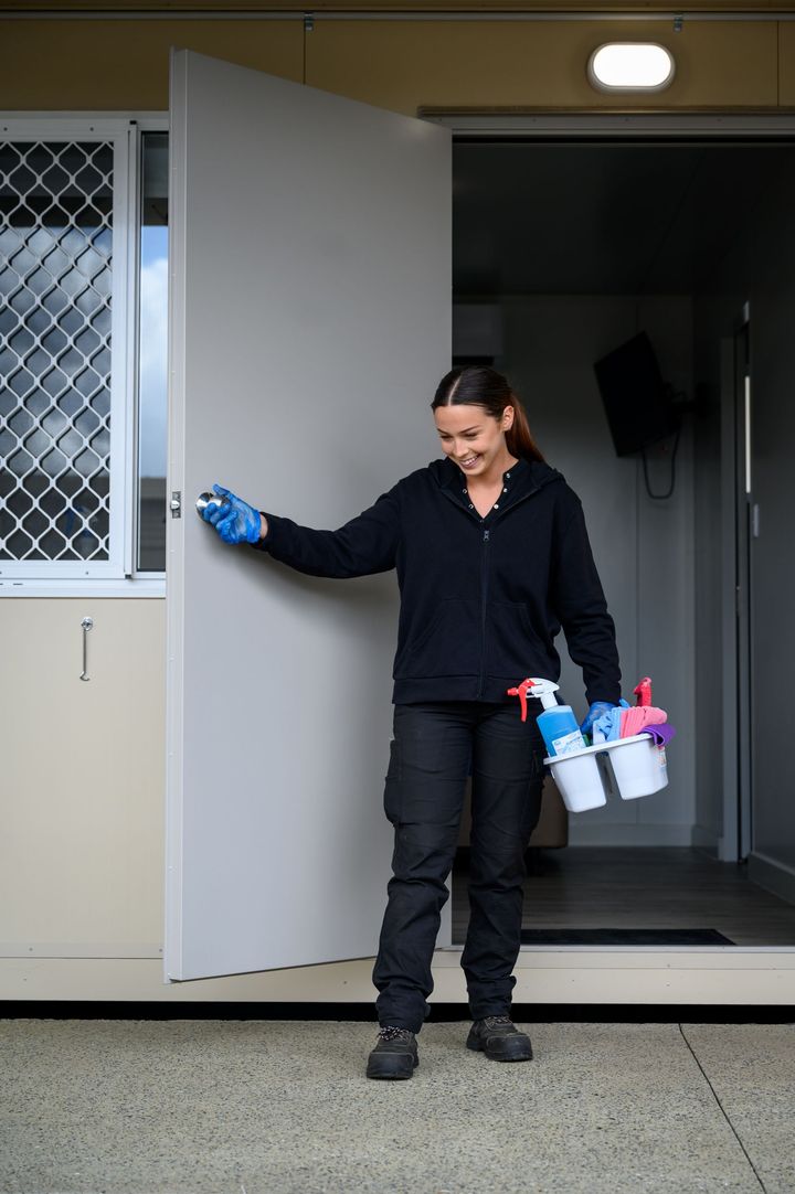 Woman in cleaning attire opening a door at Collie Hills Village, holding cleaning supplies, smiling.