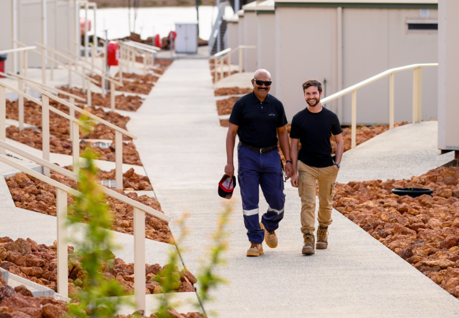 Two men walking side-by-side on a walkway between buildings in a work site setting.