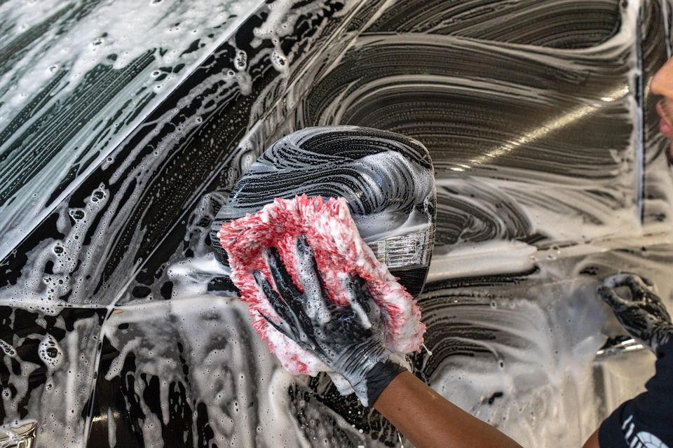 Person washing a car window with a red-and-white sponge, leaving soapy streaks behind