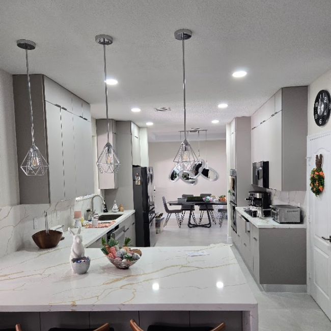 Modern kitchen with gray cabinets, marble countertops, pendant lights, and an island. Dining area visible.