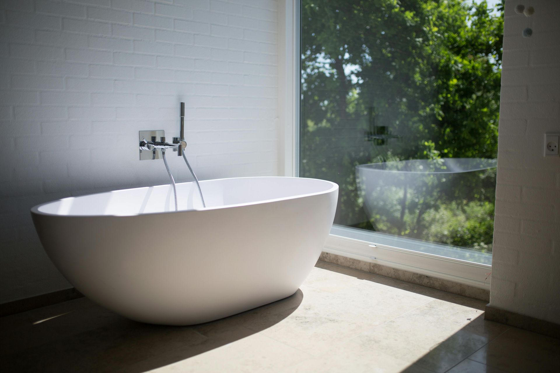 Modern white freestanding bathtub near a large window overlooking trees.