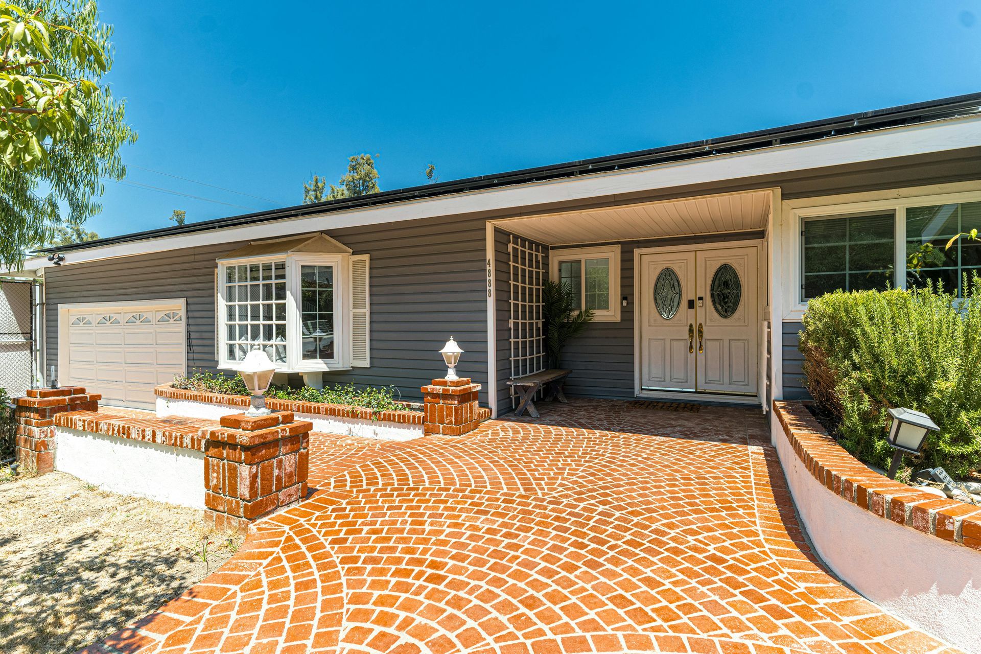 A gray ranch-style house with a red brick driveway and landscaping on a sunny day.