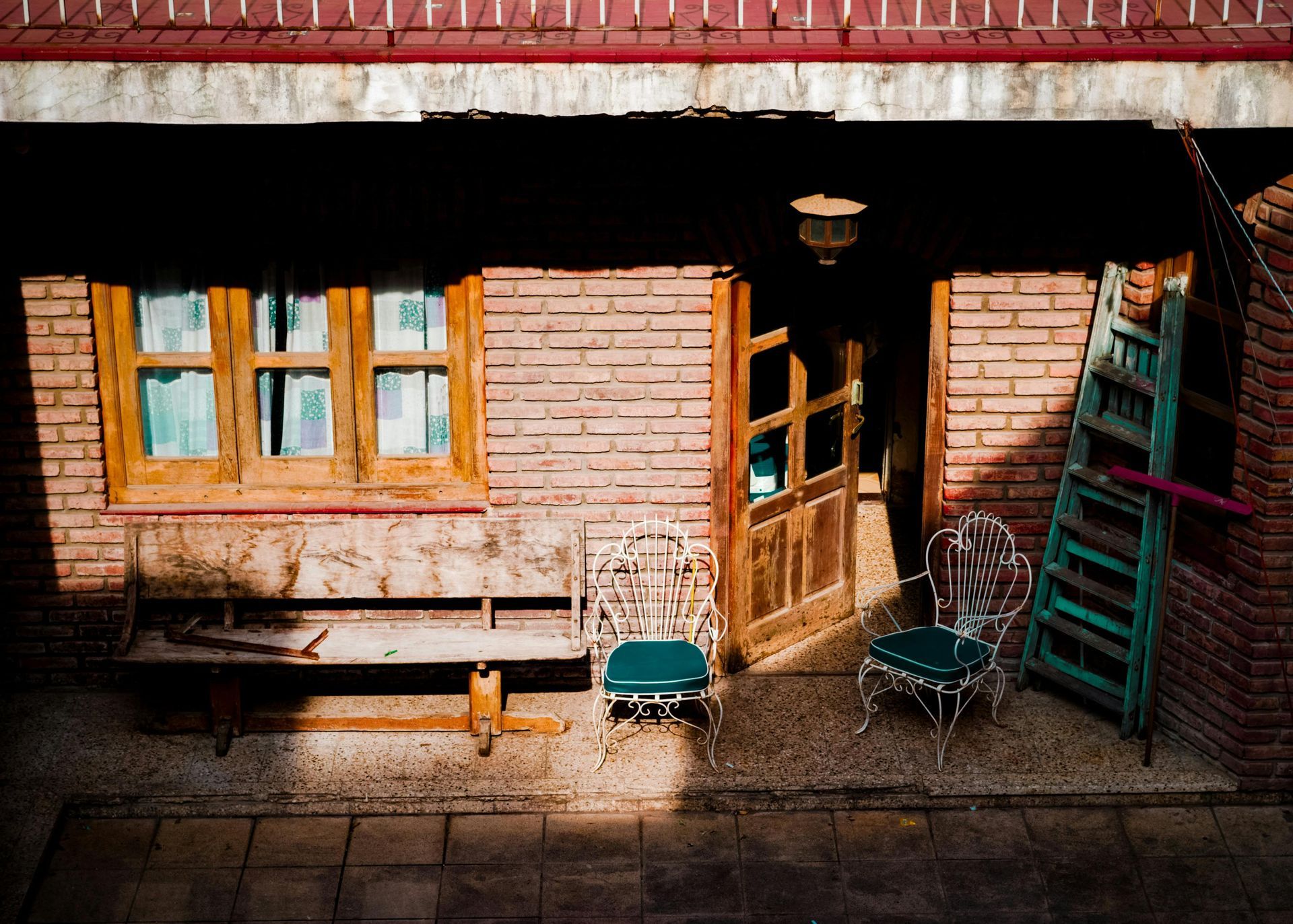 Brick building with open wooden door, window, bench, metal chairs, and ladder in sunlight.