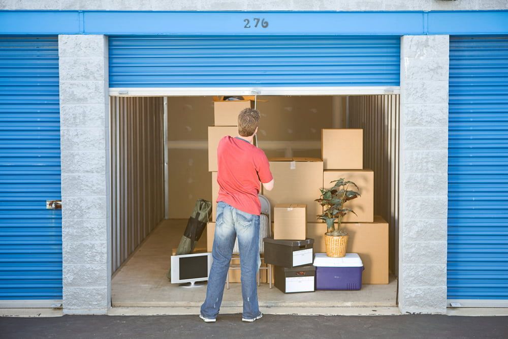 A Man In A Red Shirt Is Standing In A Storage Unit Filled With Boxes — Newcastle Moving & Storage in Salt Ash, NSW