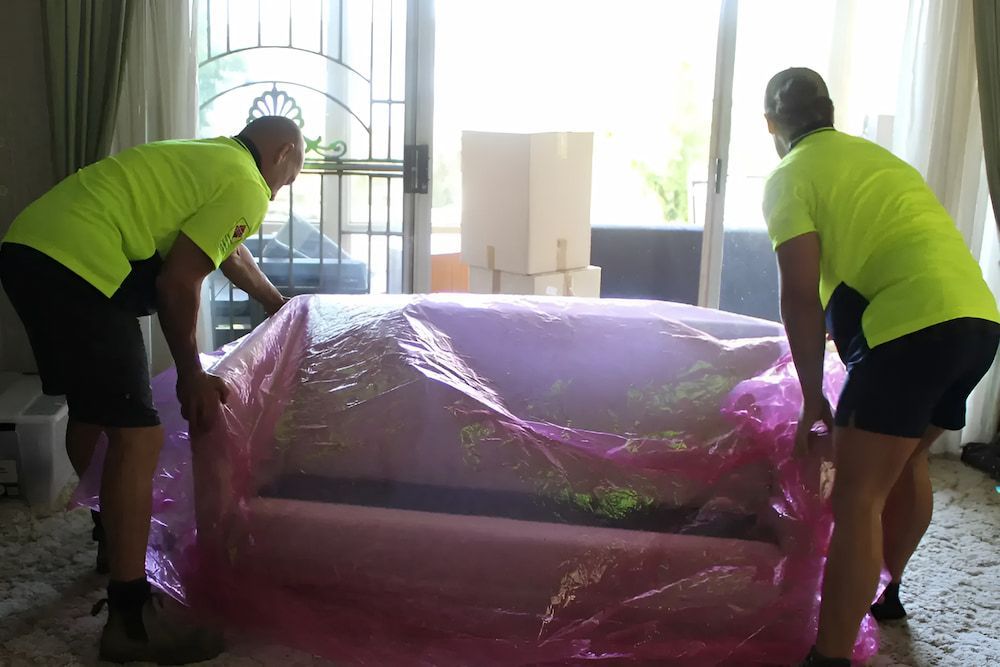 Two Men Are Wrapping A Couch In Plastic In A Living Room — Newcastle Moving & Storage in Salt Ash, NSW
