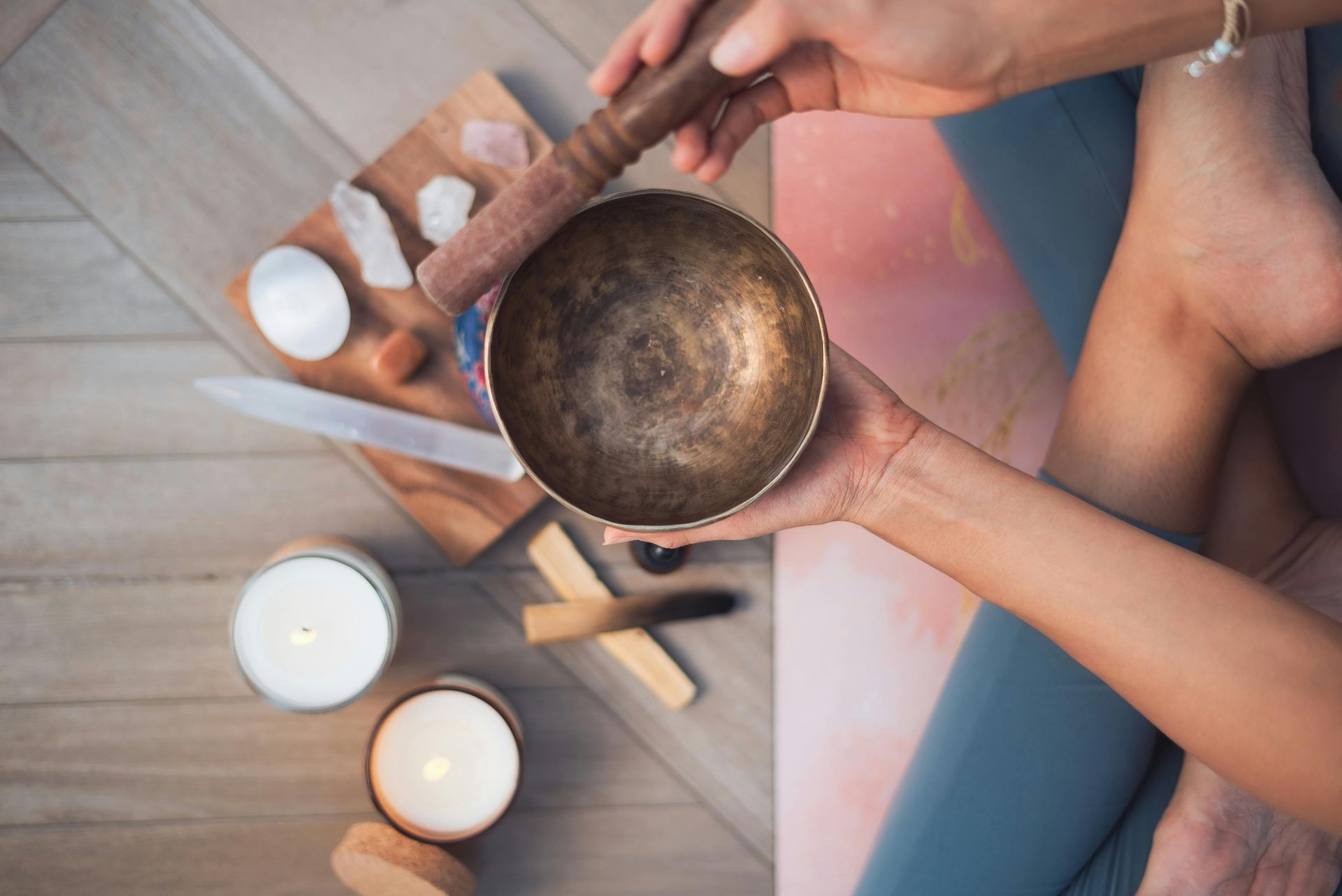 Person holding a singing bowl and mallet, with candles, crystals, and yoga mat; setting is calm.