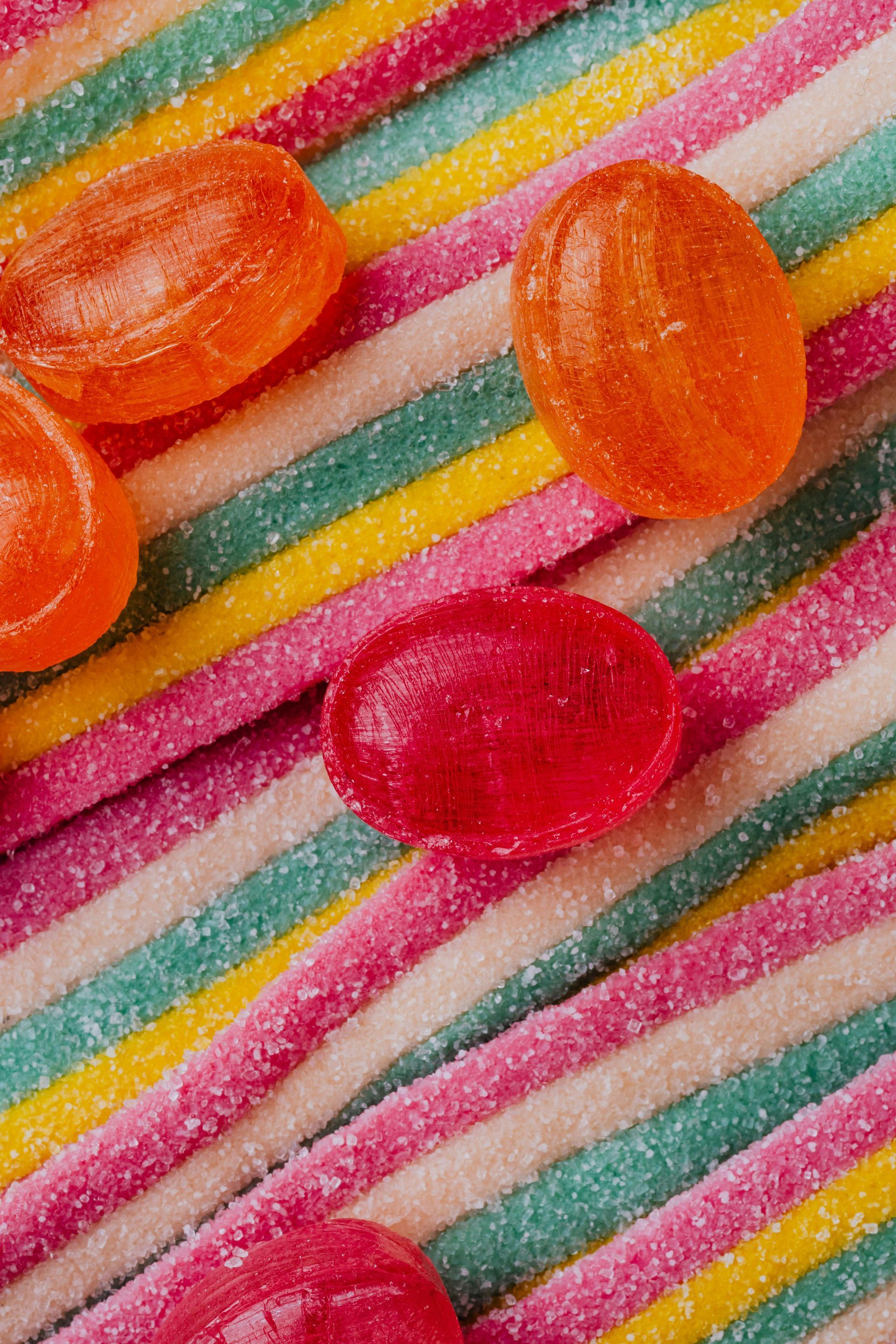 Close-up of hard candies and striped sour candy sticks, in orange and red, on a bed of green, yellow, pink, and white.