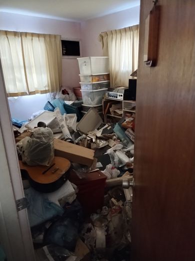 Cluttered room with piles of trash, boxes, and a guitar. Ajar door in the foreground, window with curtains in the background.