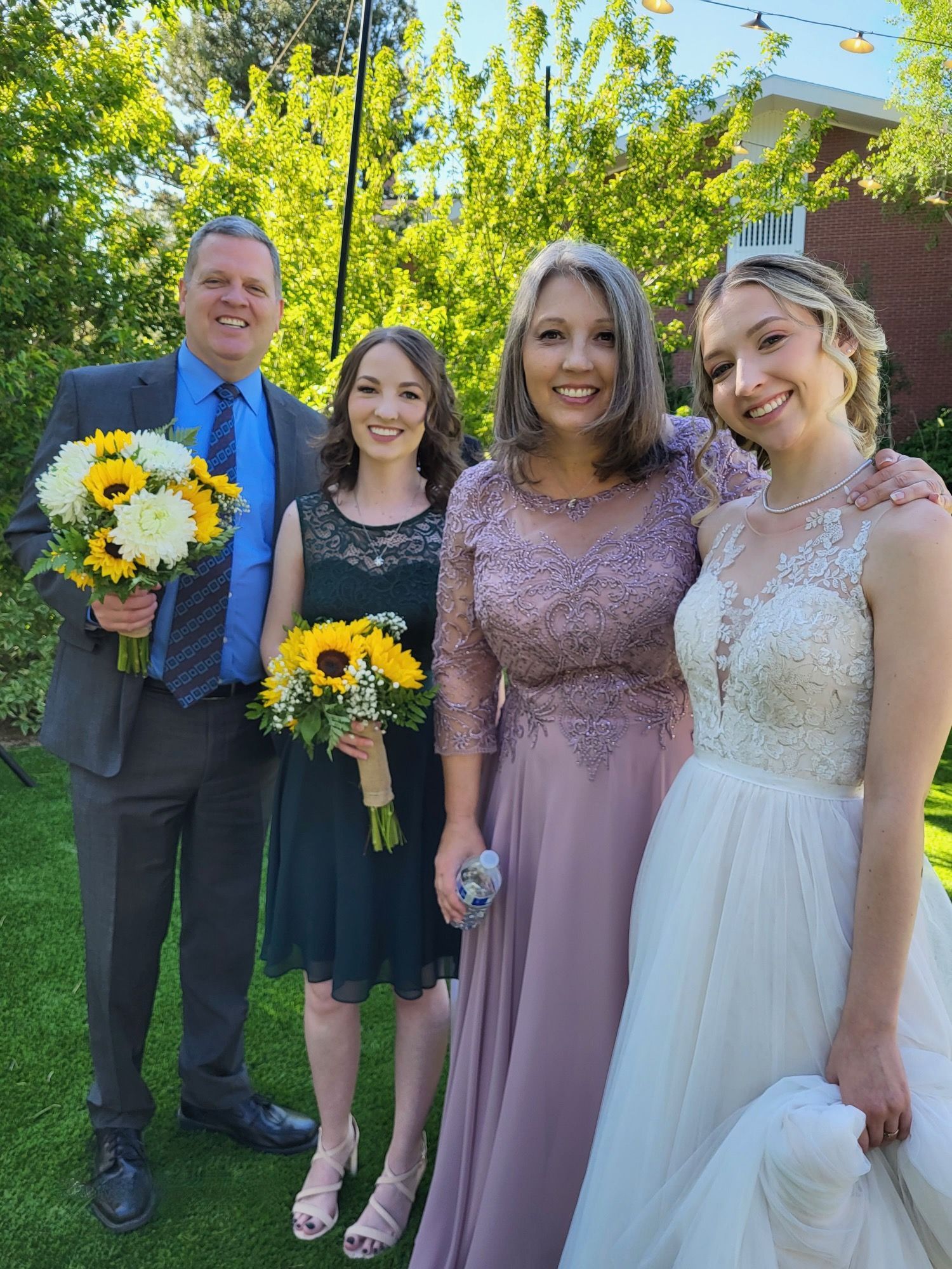 Family poses outdoors, a wedding celebration. The bride wears a white gown, and others hold flowers, smiling.