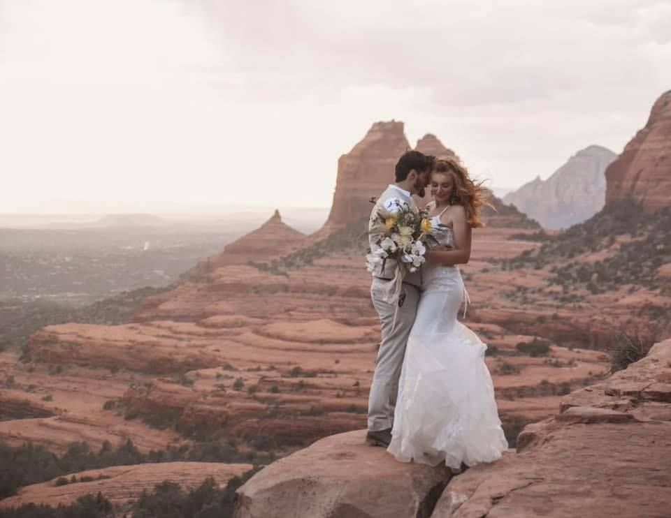 Couple embraces on a red rock cliff at sunset; the bride in a white gown, holding a bouquet.