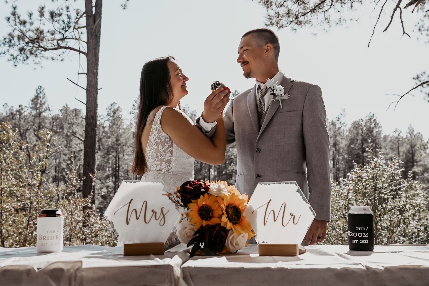 Newlyweds feed each other cake outdoors, laughing.  Sunflowers and 