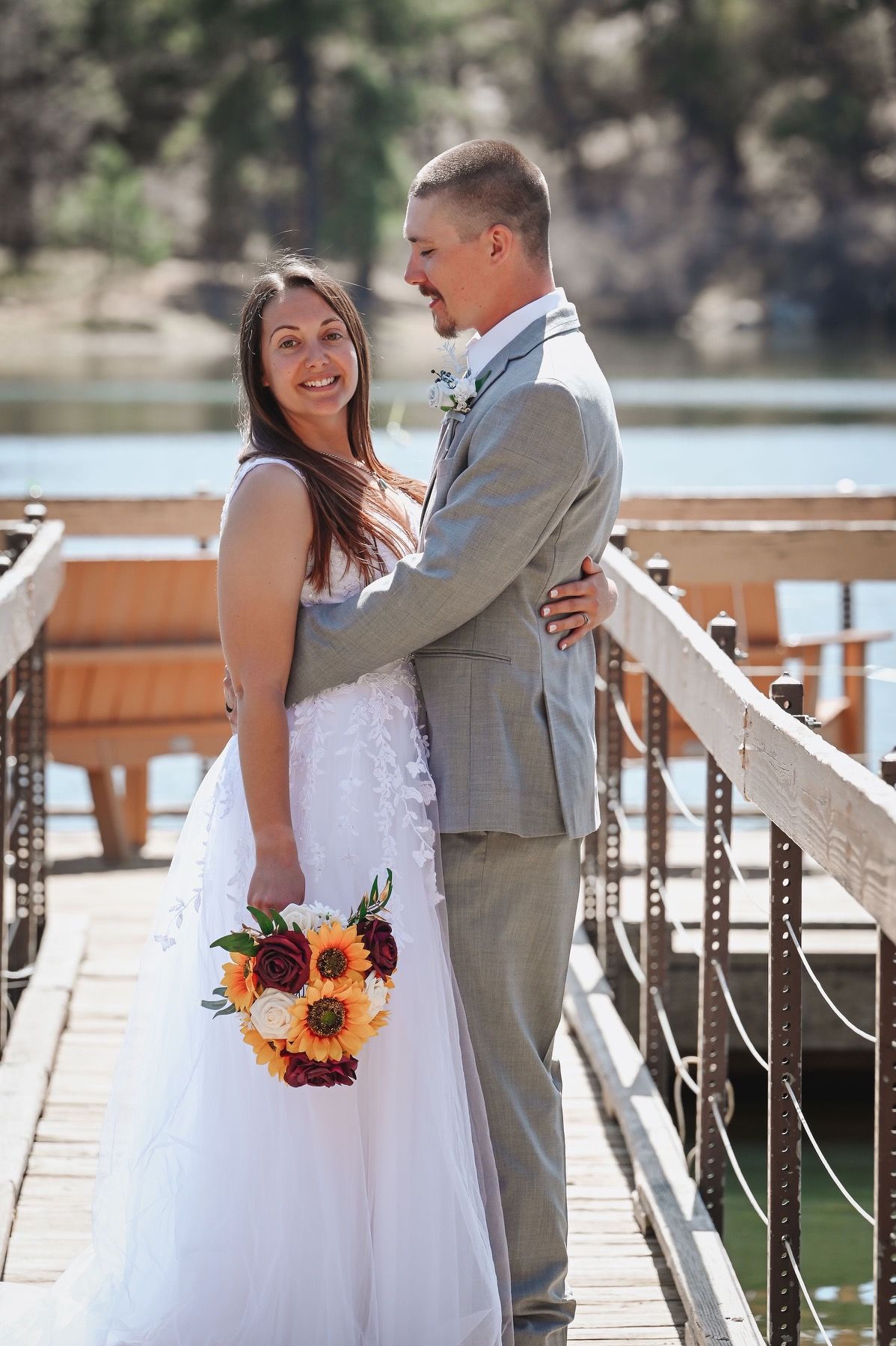 Bride and groom embrace on a wooden dock overlooking water. Bride holds a bouquet, groom wears a gray suit.
