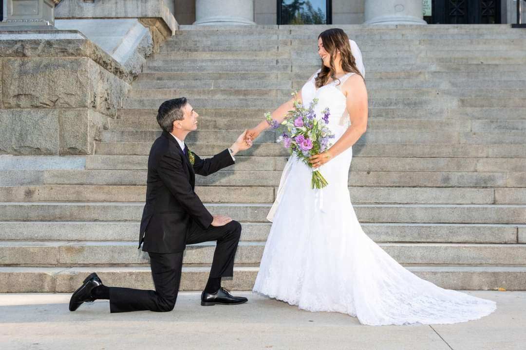 Man kneeling, proposing to woman in wedding dress on outdoor steps, holding hands.