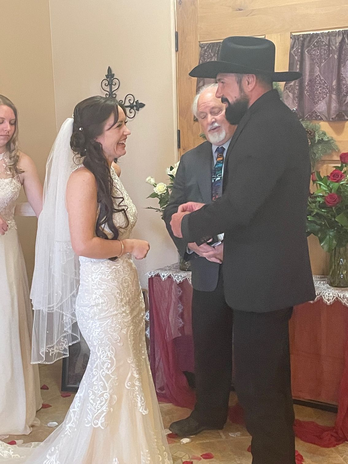 Bride and groom at altar during wedding ceremony; both smiling, officiant, bridesmaid. Room with decor.