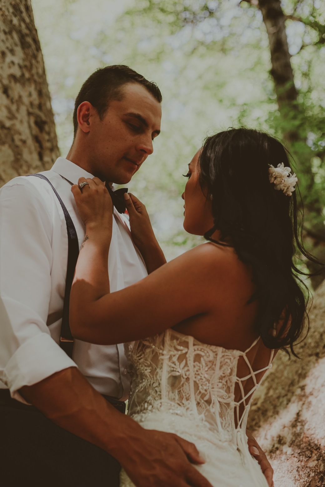 Bride adjusting groom's bow tie. They stand close together, outdoors, with trees in the background.