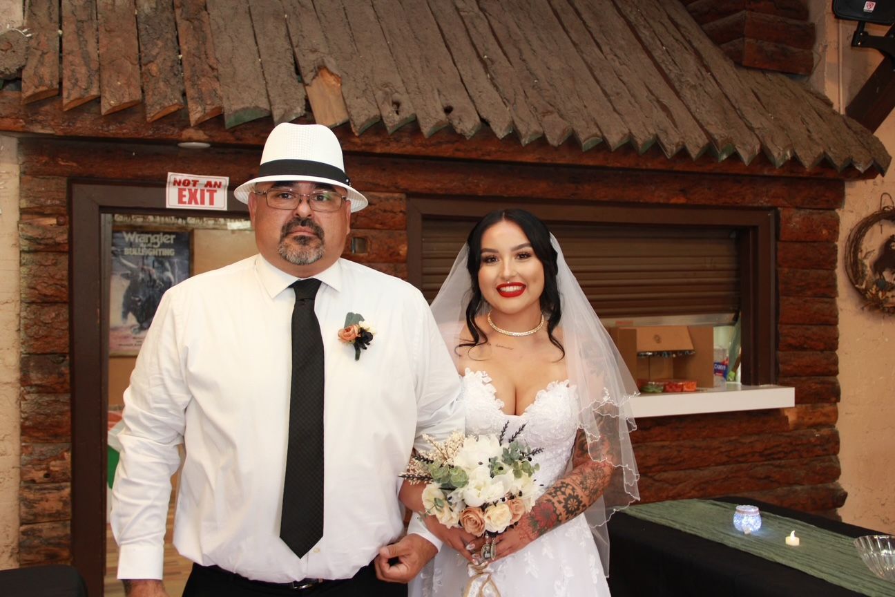 Bride in white gown and father in hat pose outside rustic building.