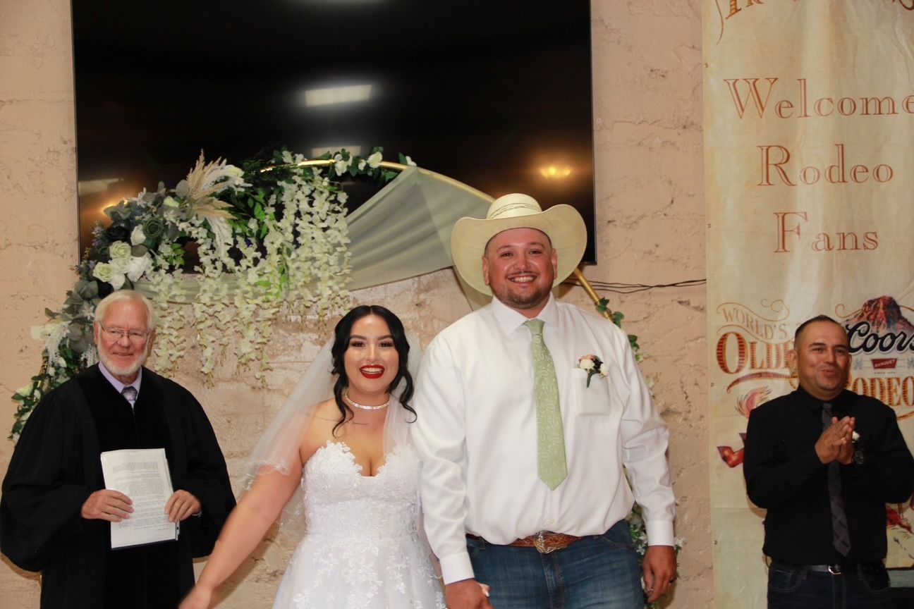 Bride and groom smiling at their wedding ceremony. The officiant and another person stand nearby. Rodeo banner in background.