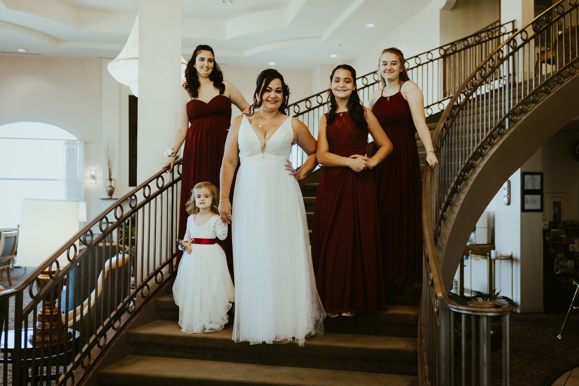 Bride in white dress descends stairs with bridesmaids in burgundy dresses, flower girl.