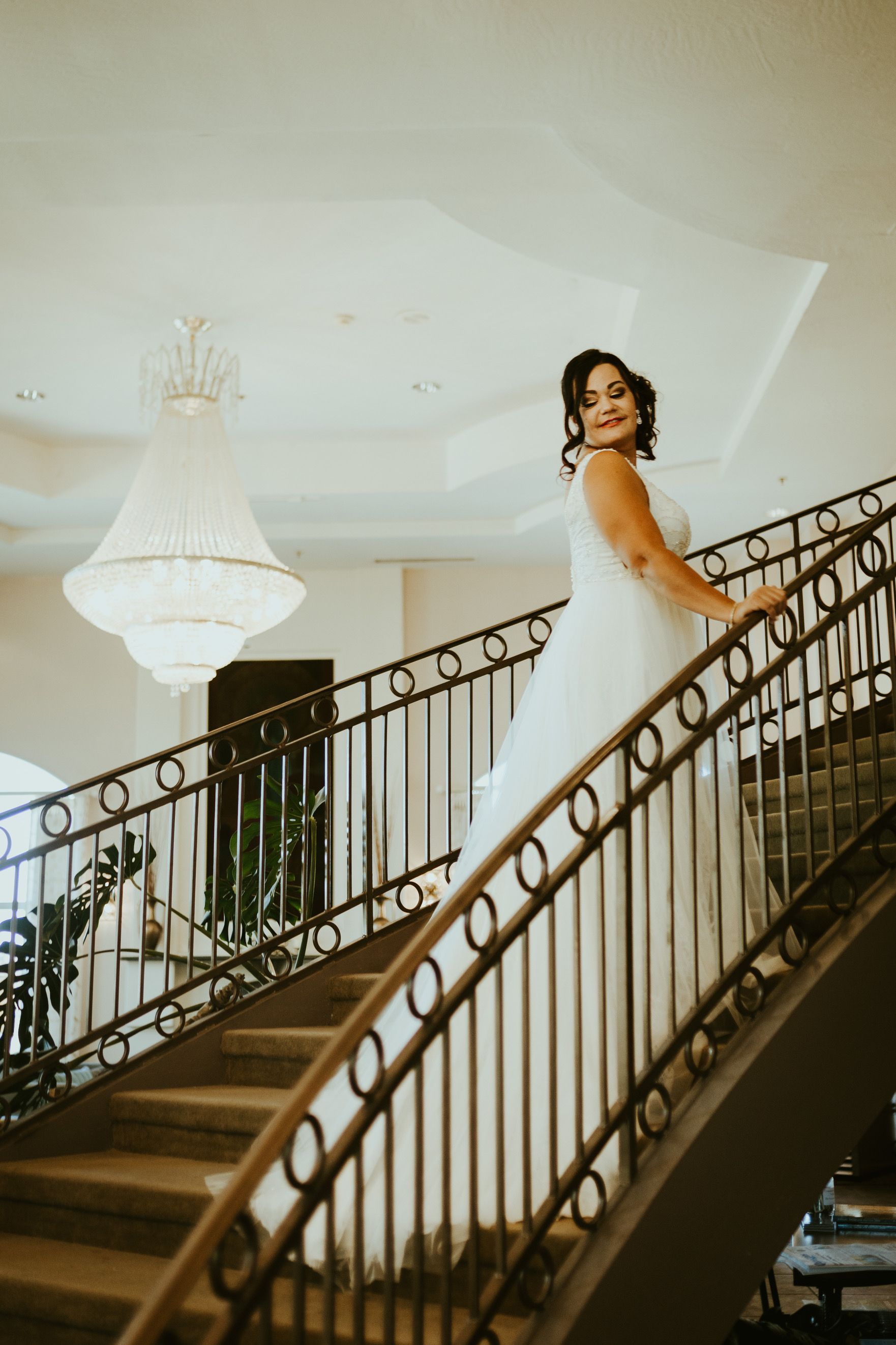 Bride in white wedding dress poses on a grand staircase, looking over shoulder, with ornate chandelier.