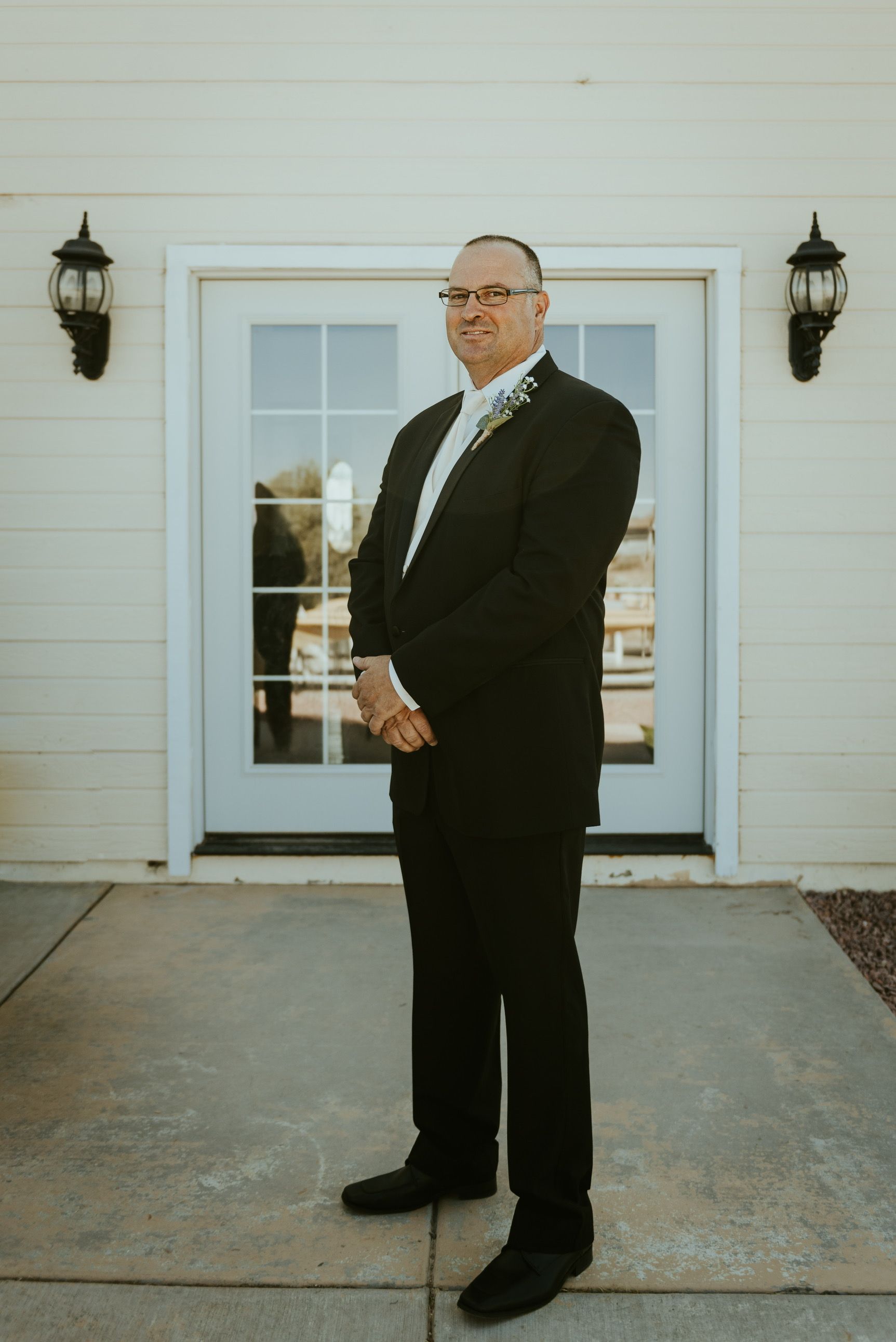 Man in a black suit stands in front of a white building with glass doors, arms crossed.