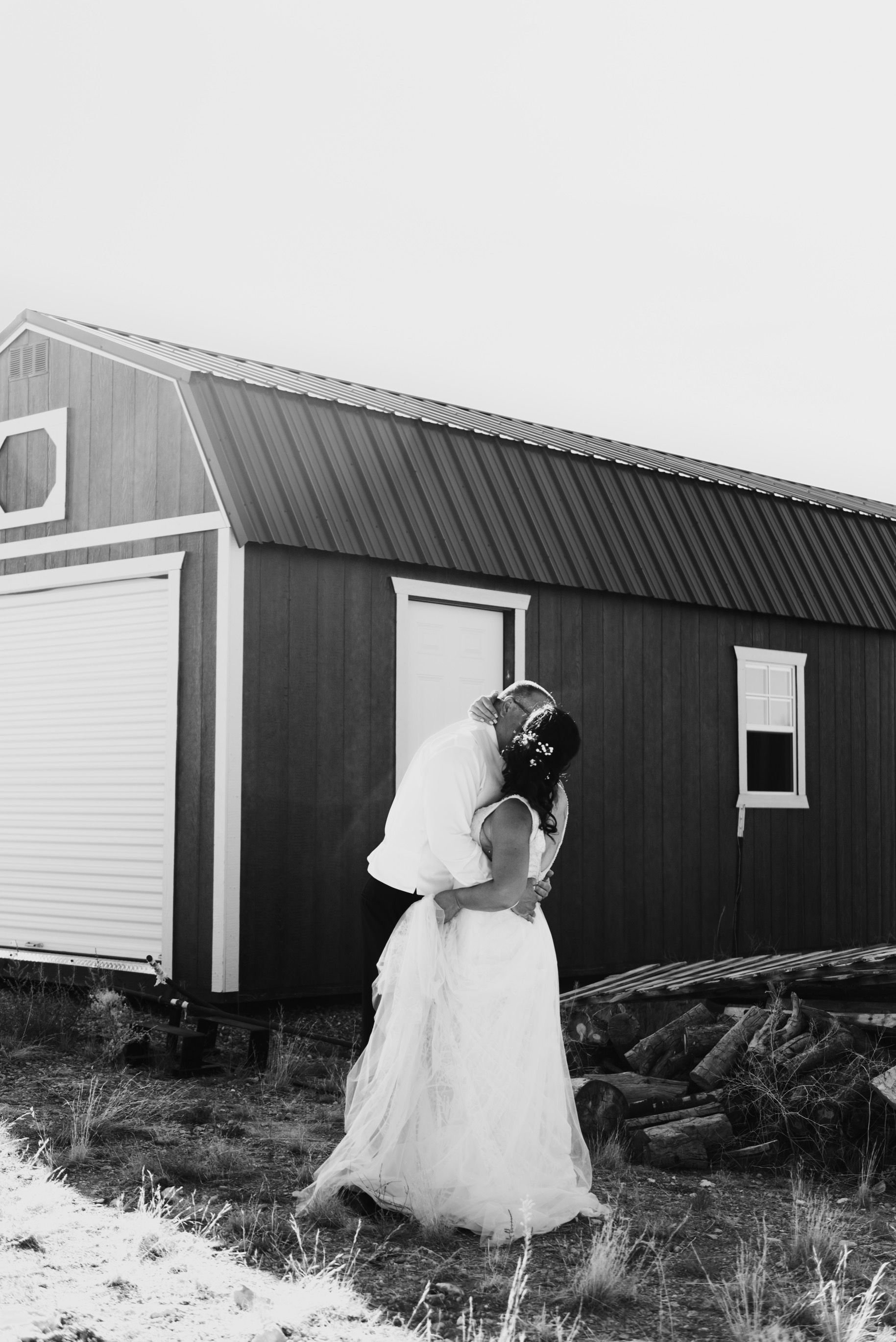 Groom lifts bride in front of a red building with white door. Both are embracing, outdoors, grayscale.