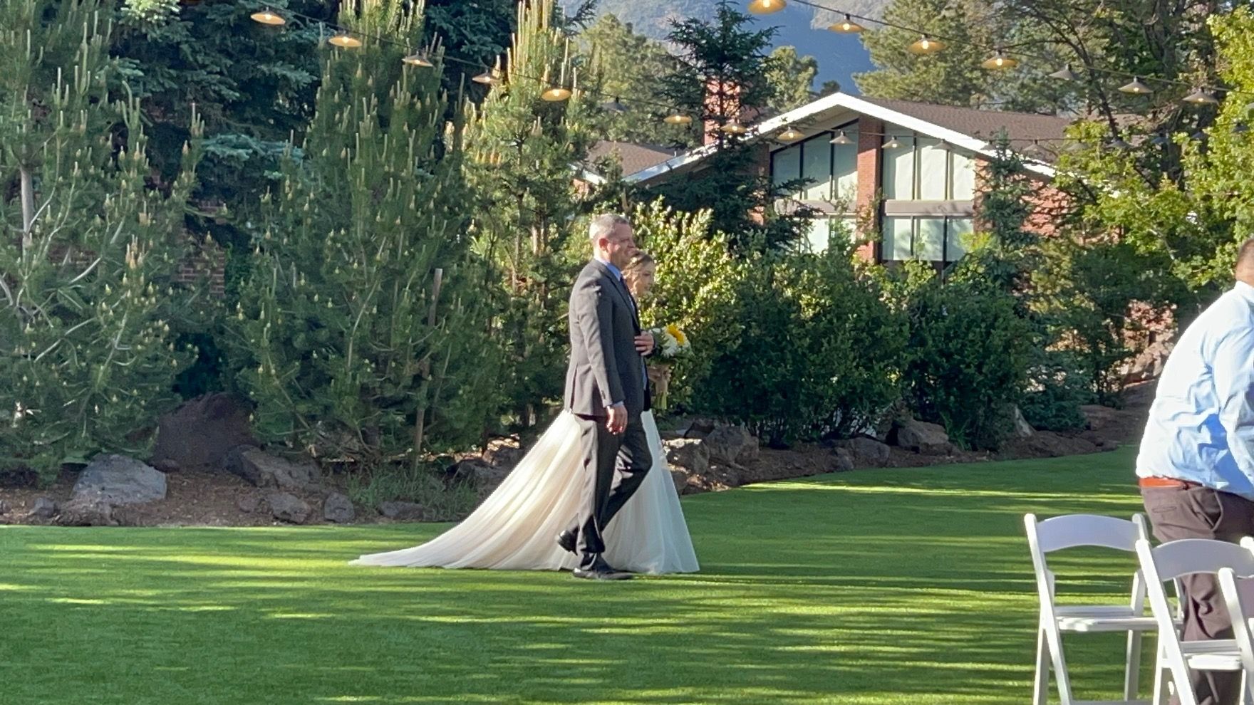 A couple walks on grass; bride in white gown, groom in gray suit. Wedding ceremony setting, house in background.