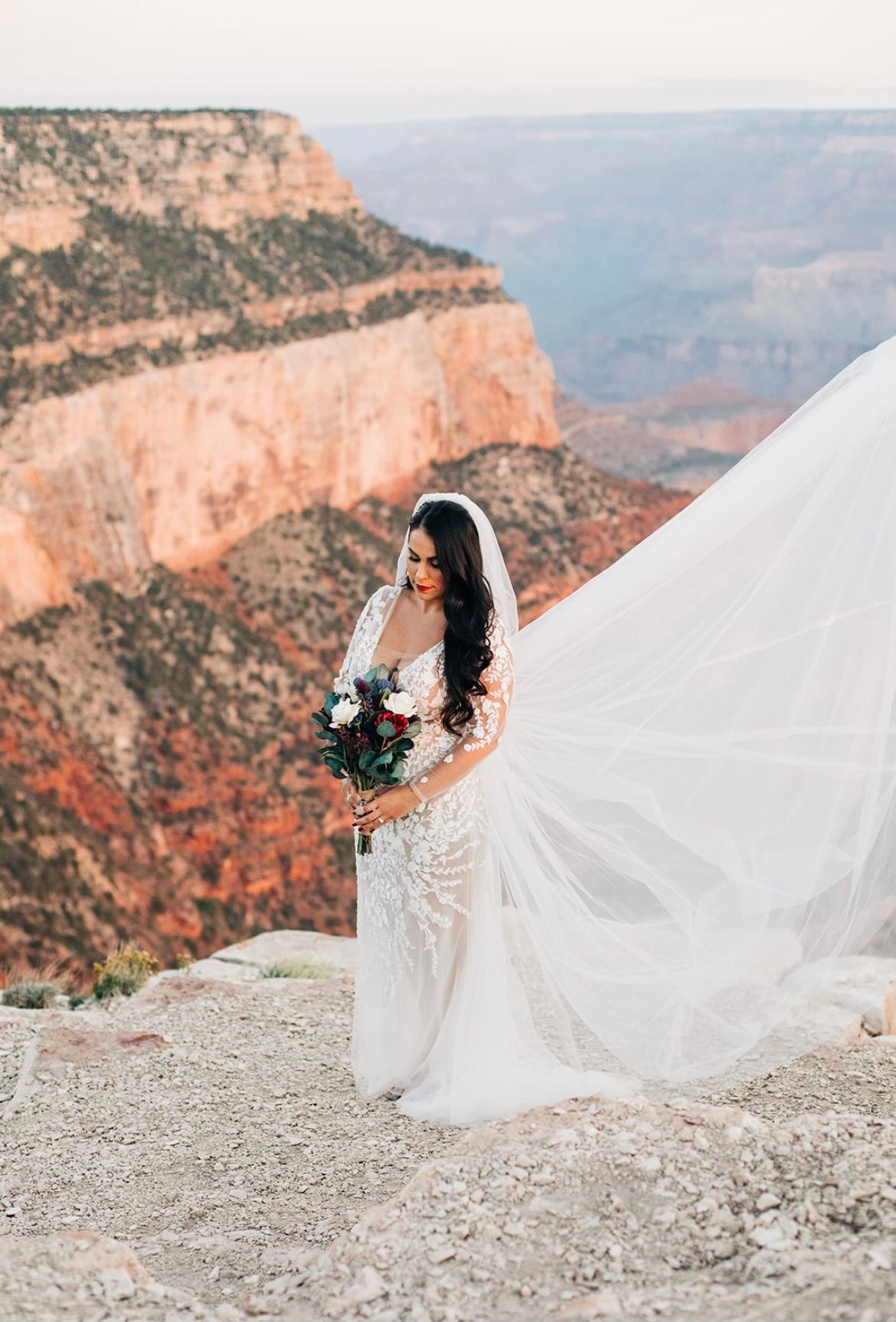 Bride in white gown holding bouquet, standing on a cliff with the Grand Canyon in the background.