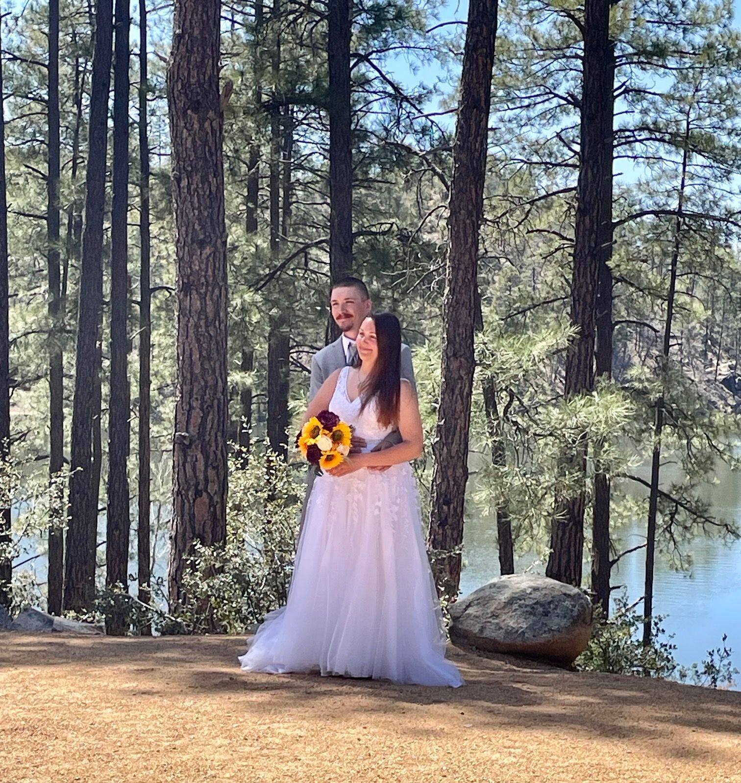 Couple in wedding attire hugging outdoors, surrounded by trees and a lake.