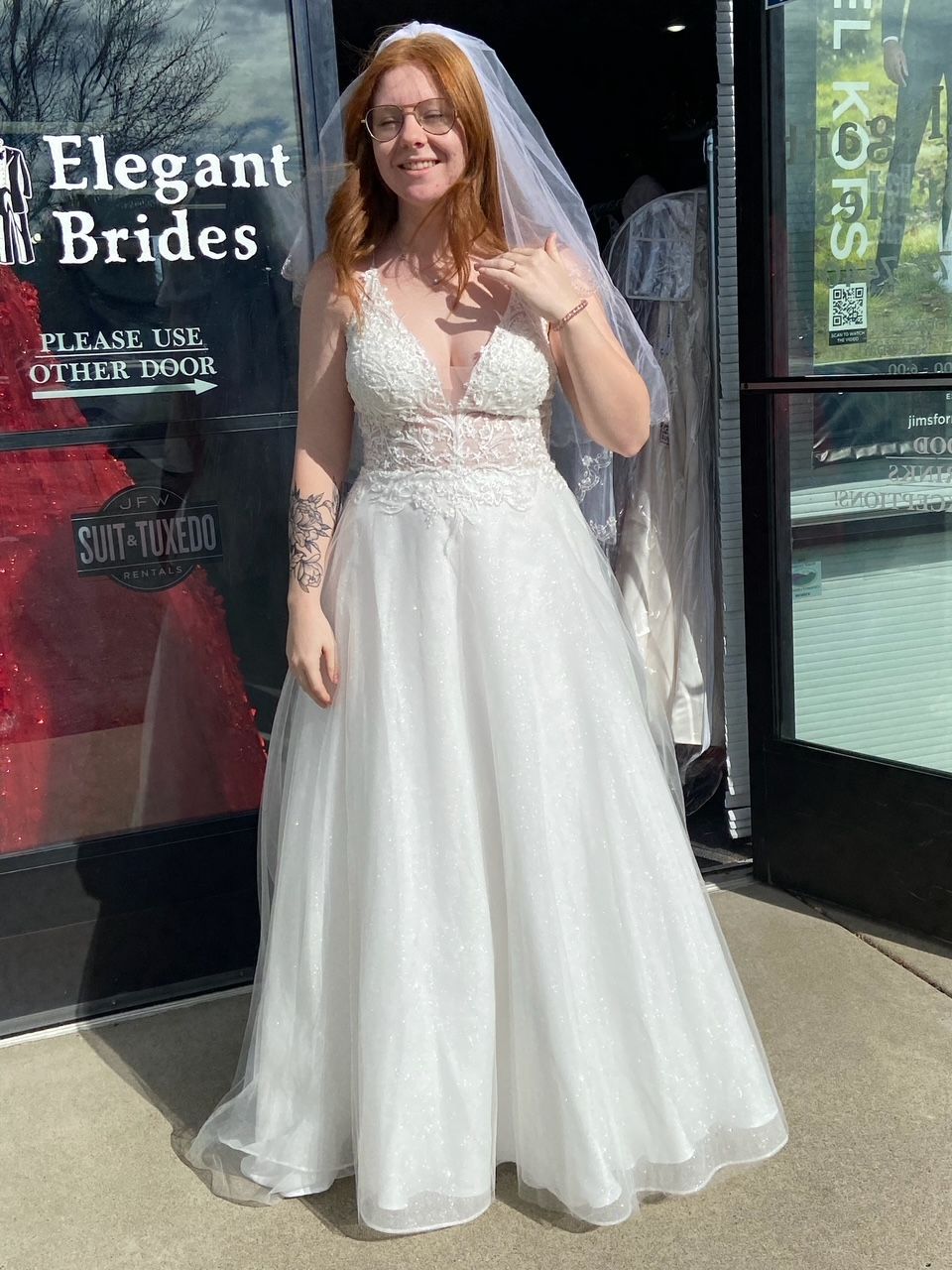 Woman in a wedding dress smiles, standing outside a bridal shop with a veil.
