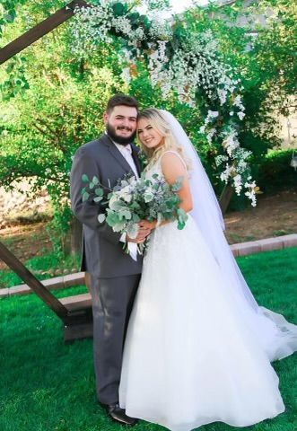 Bride and groom pose outdoors. The bride wears a white dress and veil, holding a bouquet. The groom wears a gray suit. They stand before a floral arch.