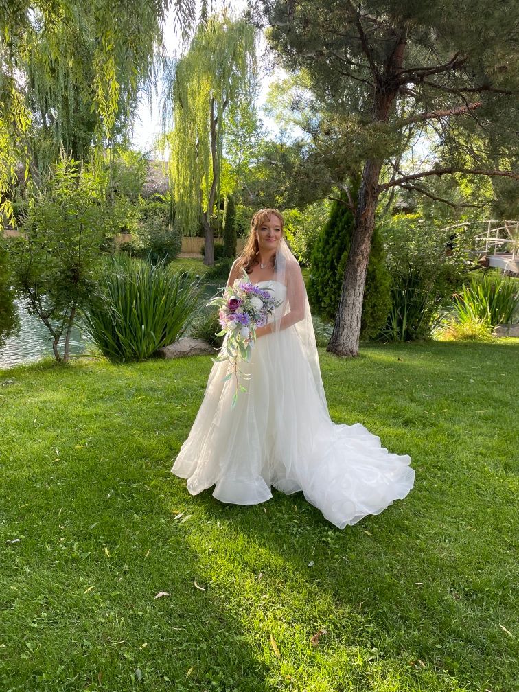 Bride in white wedding dress holding flowers, standing on green grass, outdoors.
