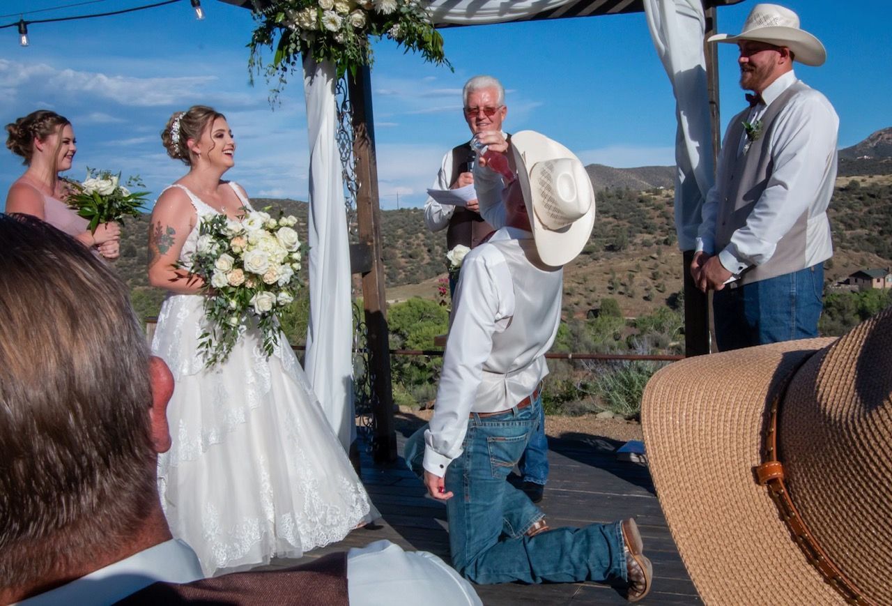 Wedding ceremony: Groom kneels, bride smiles, officiant speaks outdoors, mountainous backdrop.