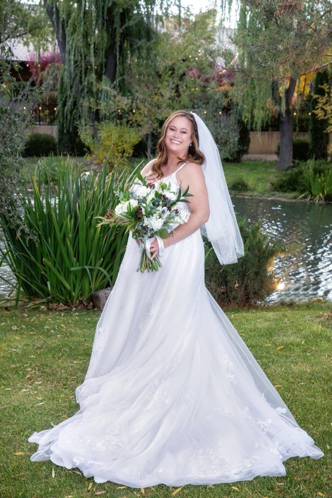 Bride in a white wedding dress holds a bouquet, standing in front of a pond and greenery.