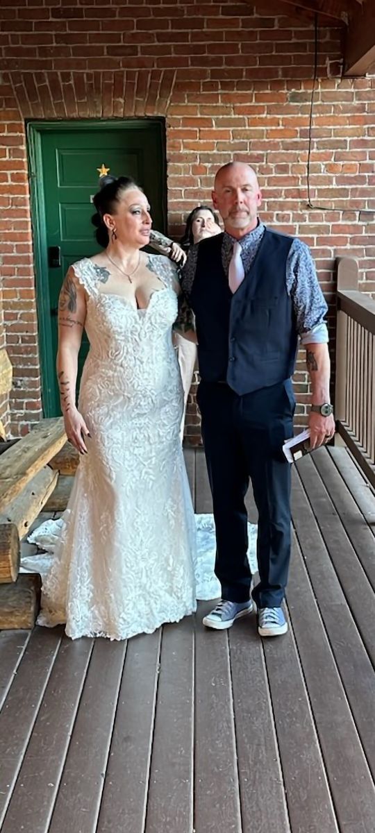 Bride and groom stand on a wooden deck in front of a brick wall and a green door. The bride wears a white dress and the groom wears a vest.